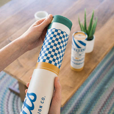 A person's hands holding a cylindrical object with a checkered pattern in blue and white colors, with a plant in a pot visible in the background.
