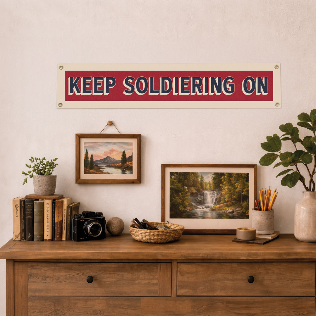 a wooden dresser with various items on top, including a framed landscape painting, a potted plant, a camera, and a sign that reads "KEEP SOLDIERING ON".