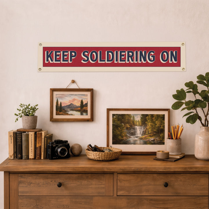 a wooden dresser with various items on top, including a framed landscape painting, a potted plant, a camera, and a sign that reads "KEEP SOLDIERING ON".