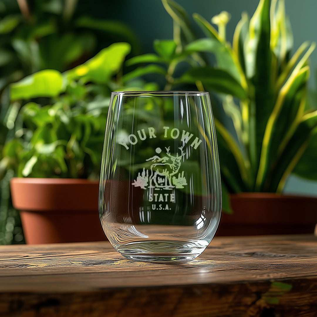 A clear glass with the text "YOUR TOWN STATE U.S.A." printed on it, placed on a wooden surface in front of potted plants.