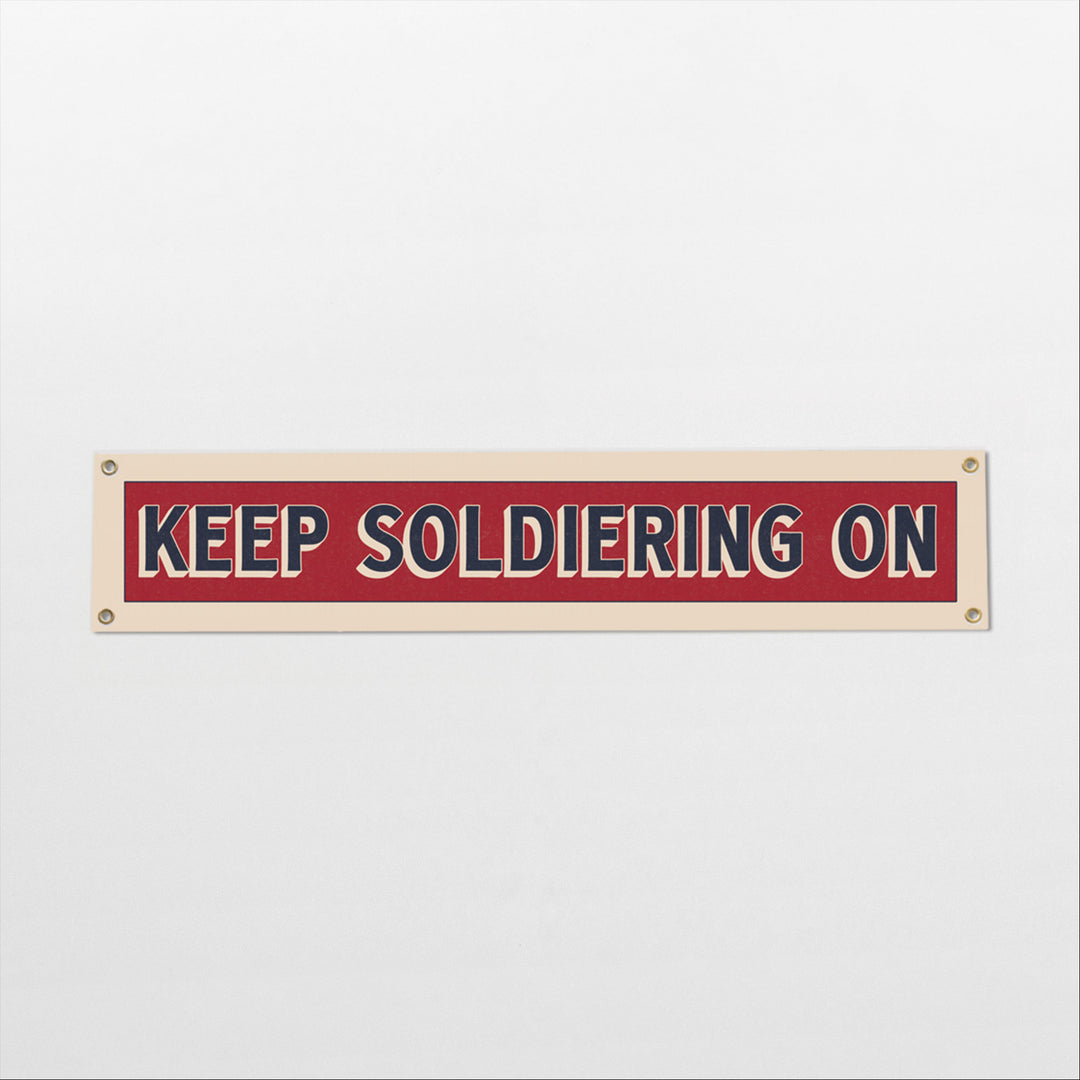 A rectangular sign with the text "KEEP SOLDIERING ON" in bold red and white letters.