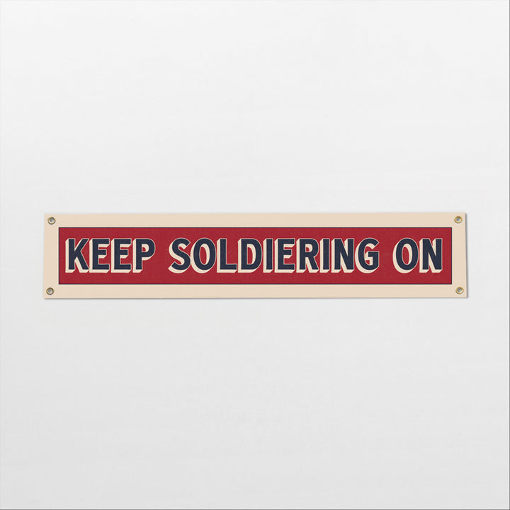A rectangular sign with the text "KEEP SOLDIERING ON" in bold red and white letters.