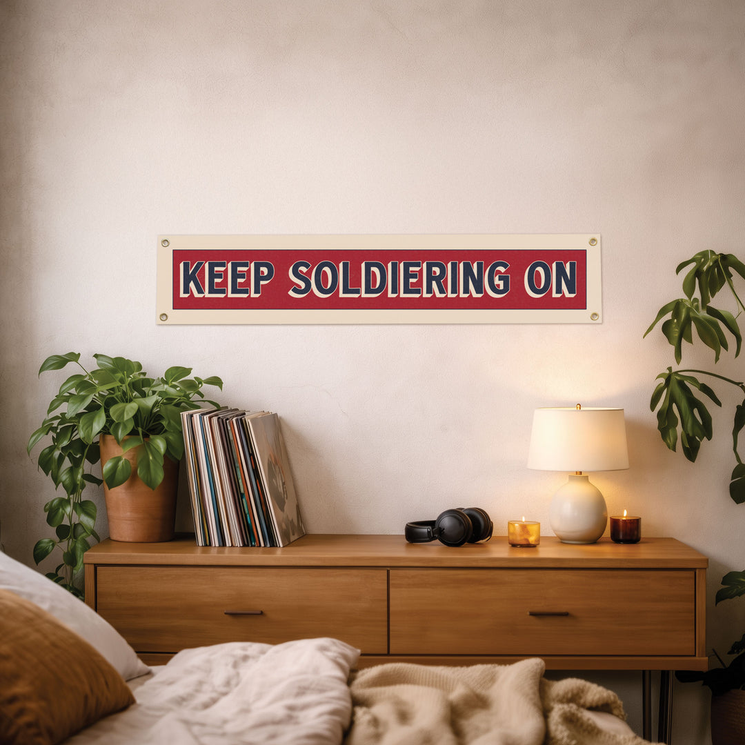 a bedroom setting with a wooden dresser, a lamp, a potted plant, and a sign that reads "KEEP SOLDIERS ON" on the wall.