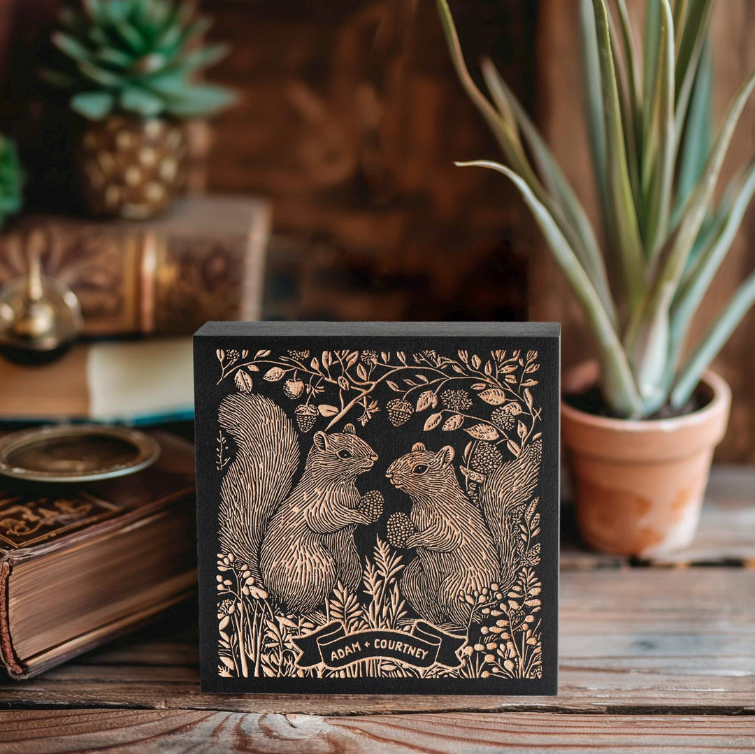 May include: A black wooden block with a gold foil design of two squirrels holding berries in a forest setting. The block has the text "Adam + Courtney" on a banner.