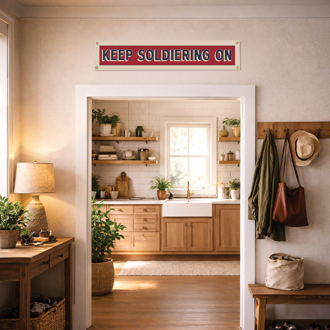 a cozy kitchen with a wooden floor and white walls, featuring a red sign that reads "KEEP SOLDIERING ON" above the kitchen sink. The kitchen is well-decorated with various potted plants and a wooden shelf.