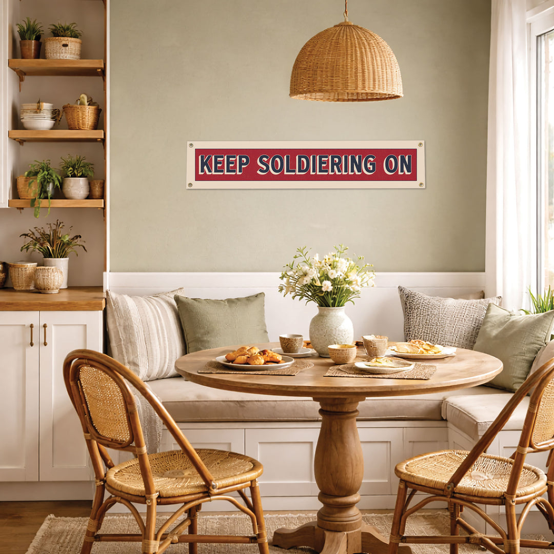 a cozy dining area with a round wooden table, two wooden chairs, and a sign that reads "KEEP SOLDIERING ON". The table is set with plates of food and a vase of flowers, creating a welcoming atmosphere.