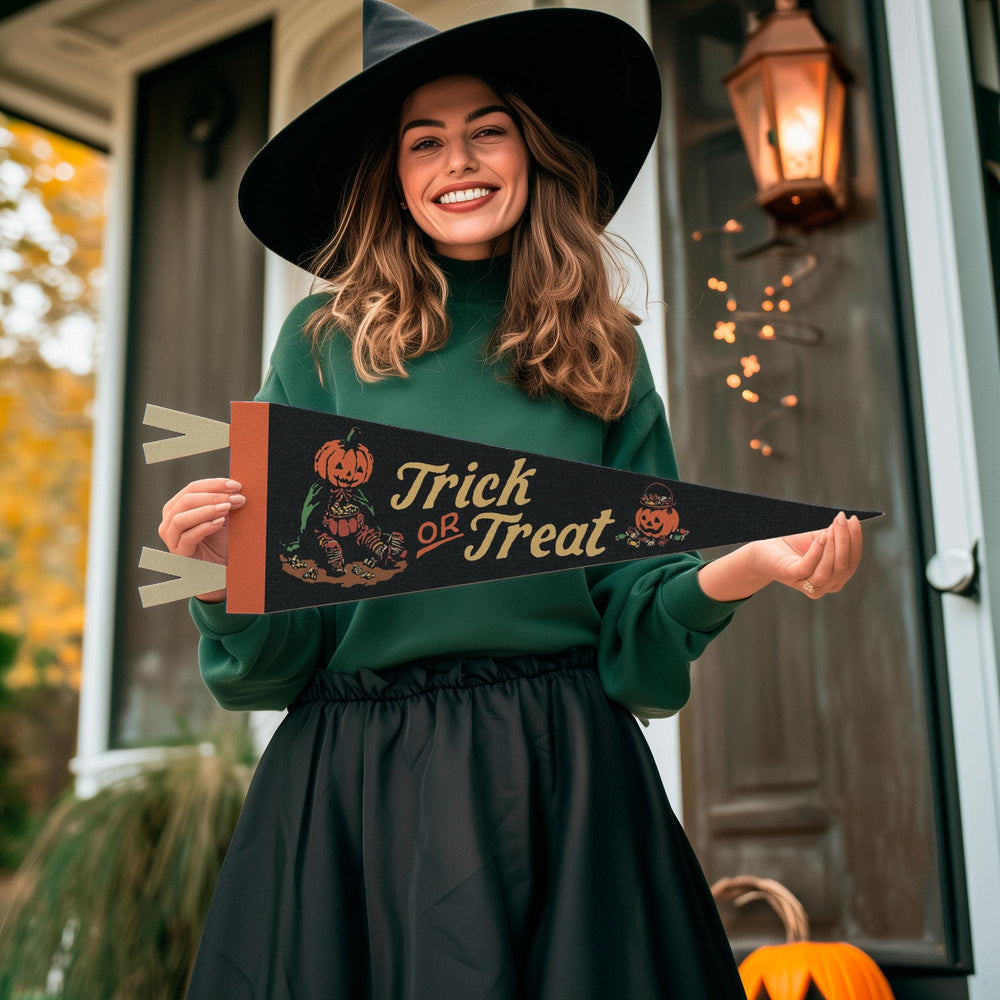 A woman wearing a black witch hat and a dark green long-sleeve top holds a black pennant banner that reads 'Trick or Treat' with jack-o'-lantern illustrations. She is standing in front of a decorated house porch with a glowing lantern and pumpkins.