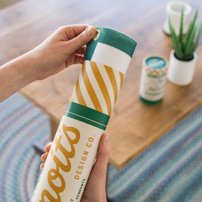 A person's hands holding a cylindrical object with a striped pattern, which appears to be a cylindrical container or tube. The background includes a wooden surface and some plants, creating a natural and indoor setting.