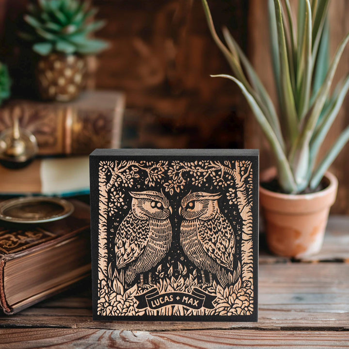 May include: A black wooden block with a gold-colored design of two owls facing each other in a forest setting. The owls have detailed feathers and large eyes. The text &amp;quot;Lucas + Max&amp;quot; is on a banner below the owls.