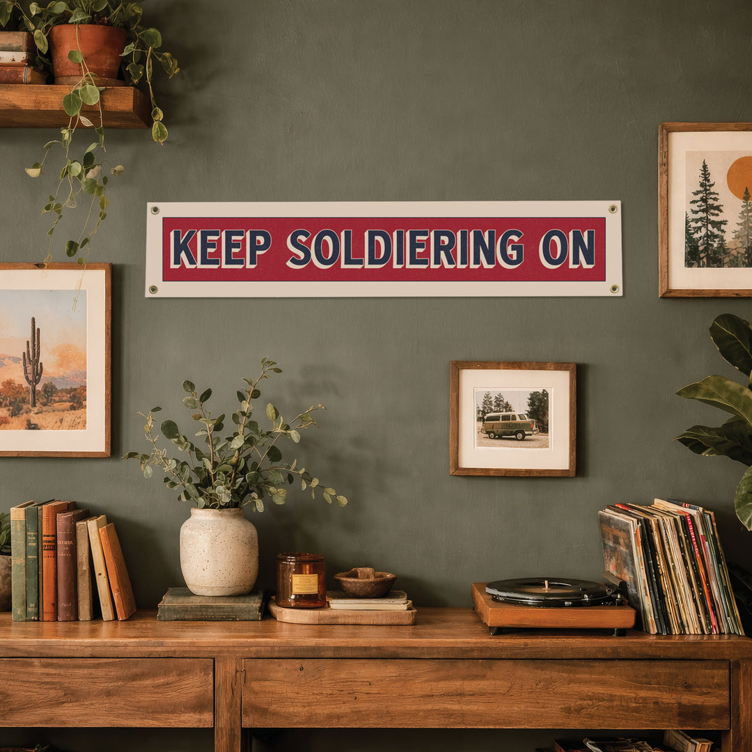 a wall with a red and white sign that says "KEEP SOLDIERING ON", surrounded by various decorative elements such as plants, books, and framed pictures.