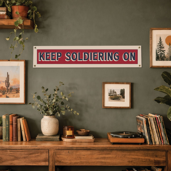a wall with a red and white sign that says "KEEP SOLDIERING ON", surrounded by various decorative elements such as plants, books, and framed pictures.
